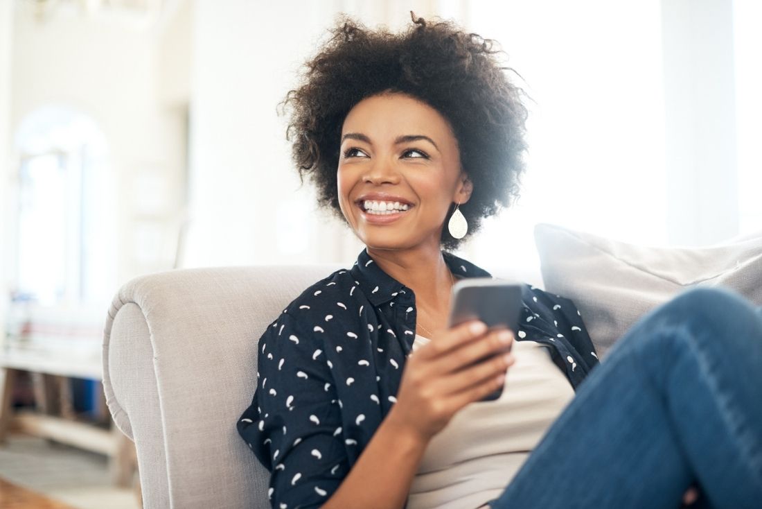 A smiling woman holding a smartphone sits comfortably on a couch in a bright, well-lit living room.