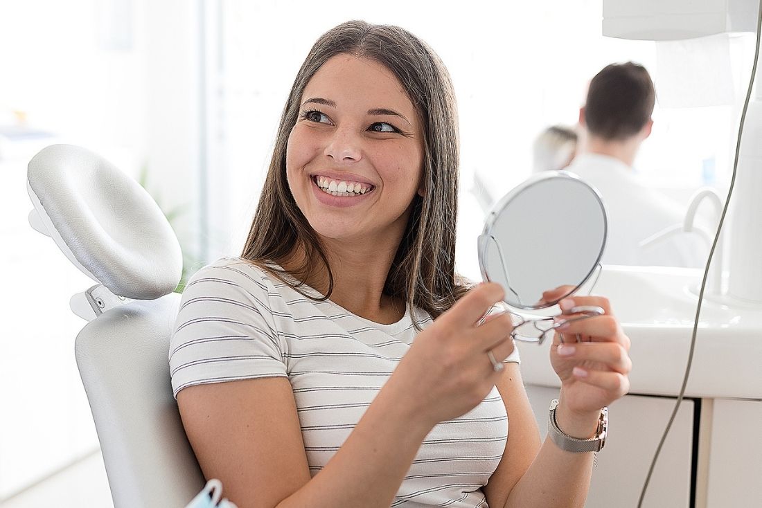 A patient smiling while holding a small mirror in a bright dental office.