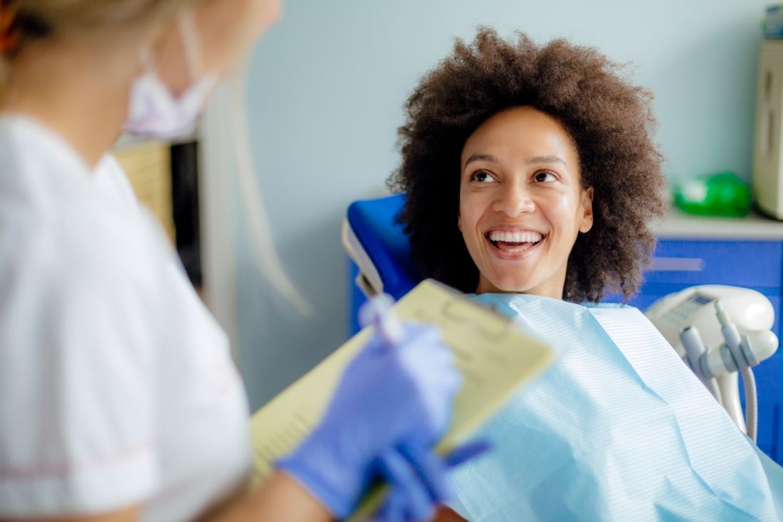 A patient smiling in a dental chair while a dental staff member reviews a clipboard in a treatment room.