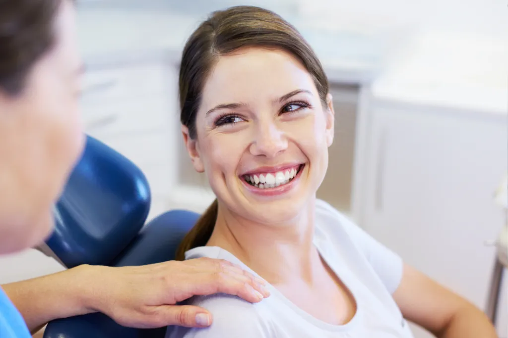 A patient smiling while sitting in a dentist's chair, with a dental professional reaching out to her in a treatment room.
