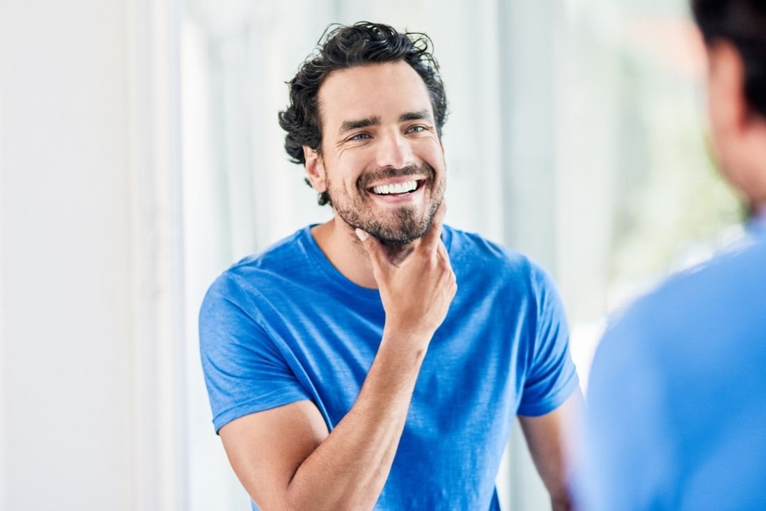 A man smiling and touching his chin while looking at his reflection in a mirror.