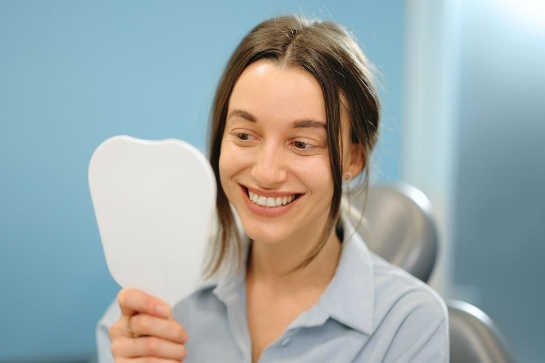 A young woman with long hair smiles while holding a small mirror in a dental office.