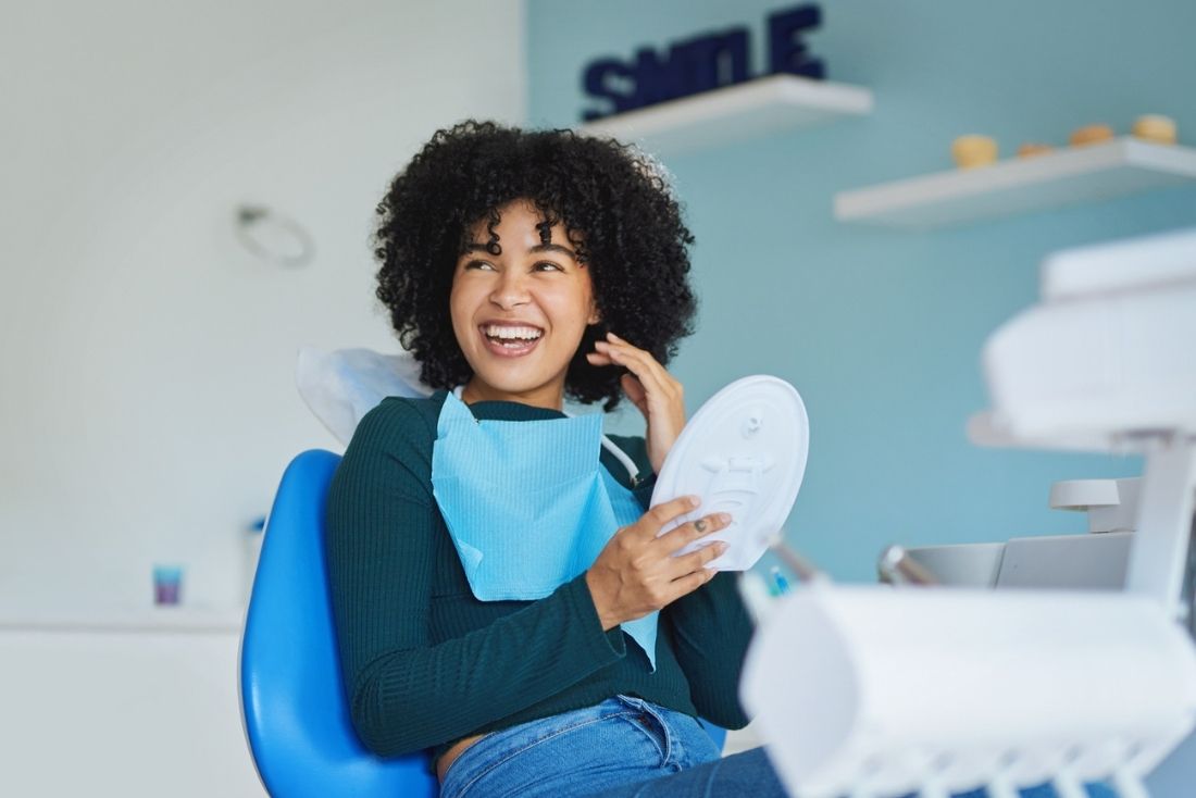 A smiling patient sitting in a dental chair with a dental bib, holding a dental tool in a bright treatment room.