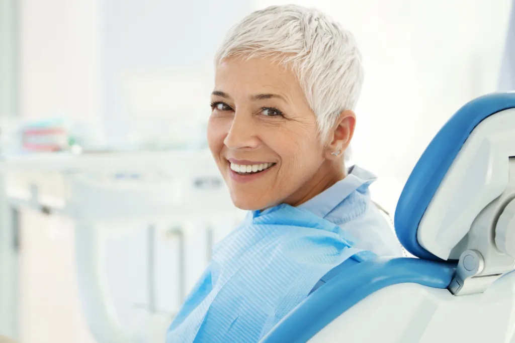 A smiling woman in a dental treatment chair wears a blue bib in a bright dental office.