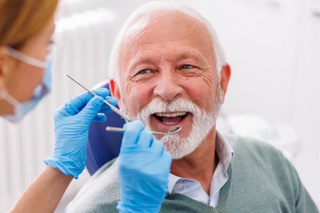 A dentist is examining an elderly man's teeth in a dental treatment room, with the man smiling.