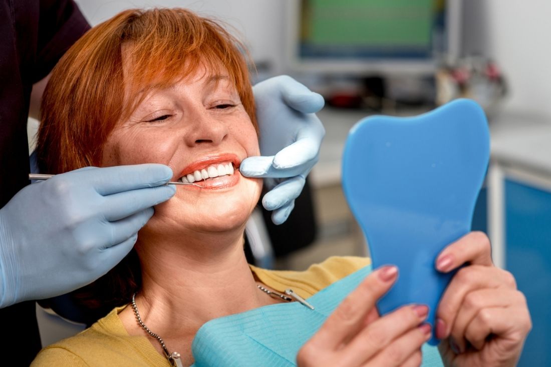 A woman in a dental chair smiles while a dentist inspects her teeth and she holds a mirror.