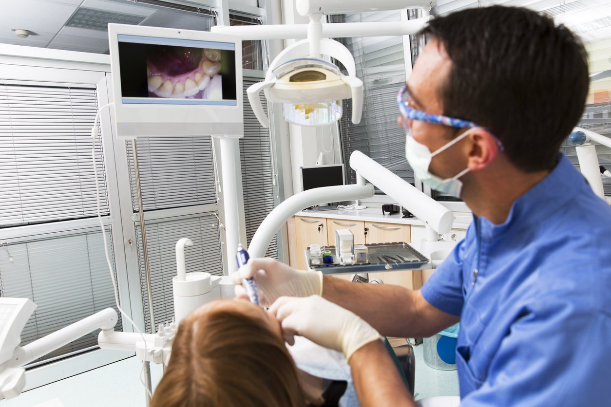 A dentist examining a patient's teeth while a monitor displays an image of the patient's mouth in a treatment room.