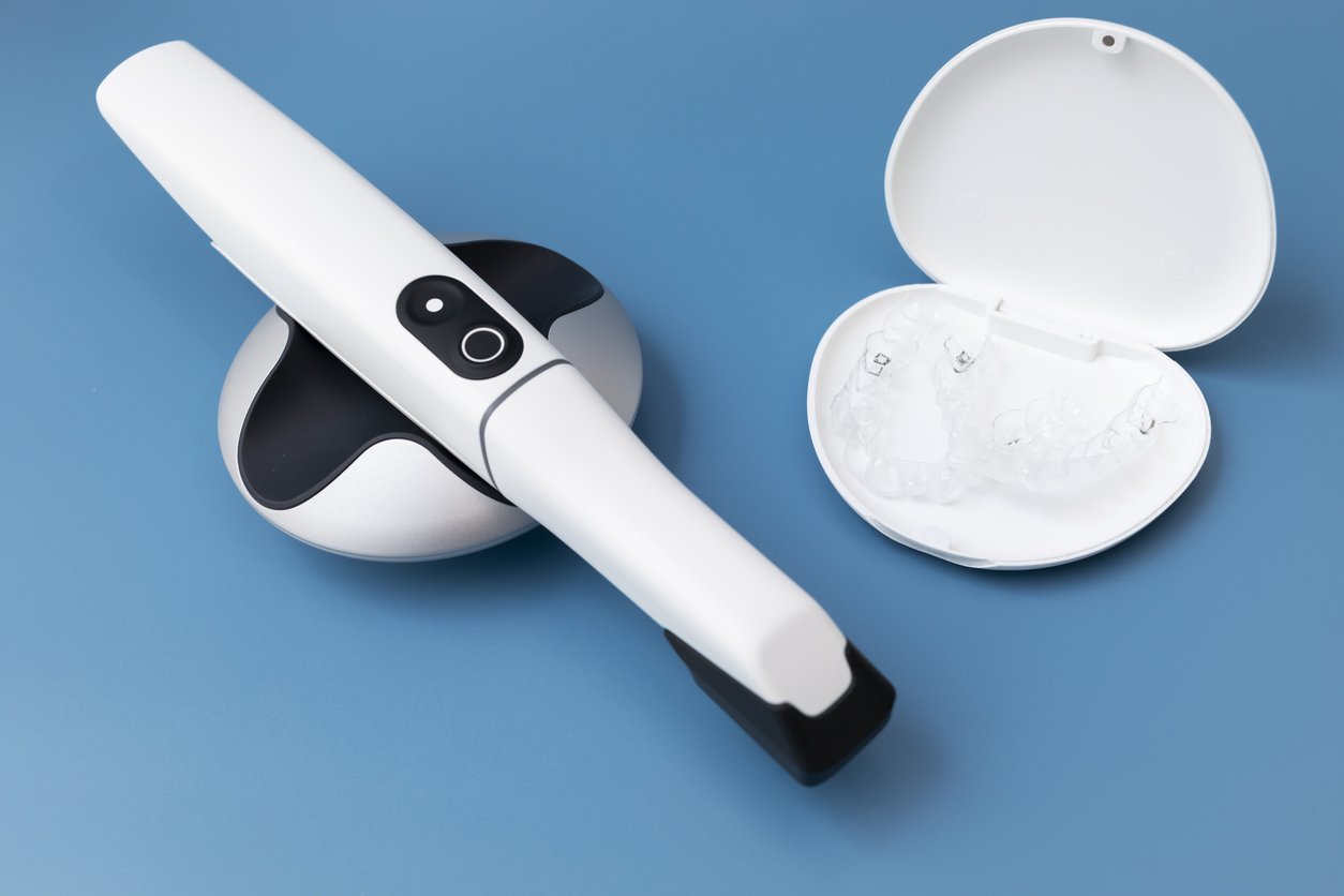 A dental scanner resting on a holder next to an open case containing clear dental aligners on a blue surface.