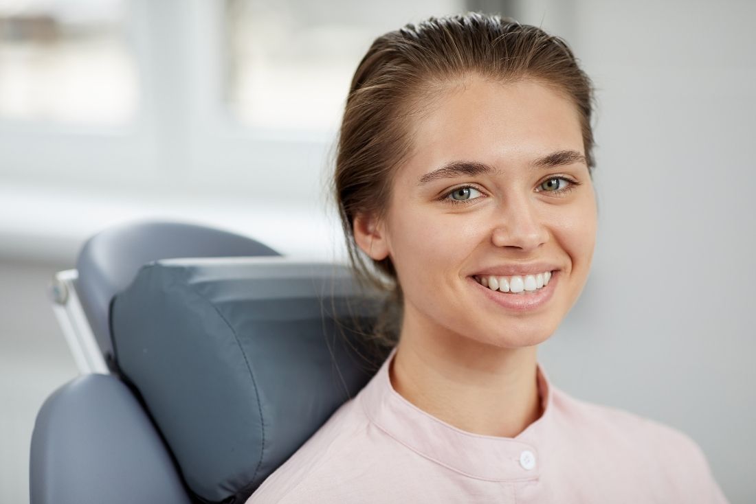 A smiling young woman in a dental office wearing a pink dental uniform and sitting in a dental chair.