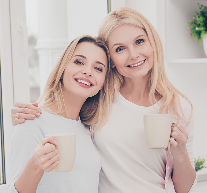 Two smiling women hold coffee mugs and pose closely together in a bright, modern room with plants.