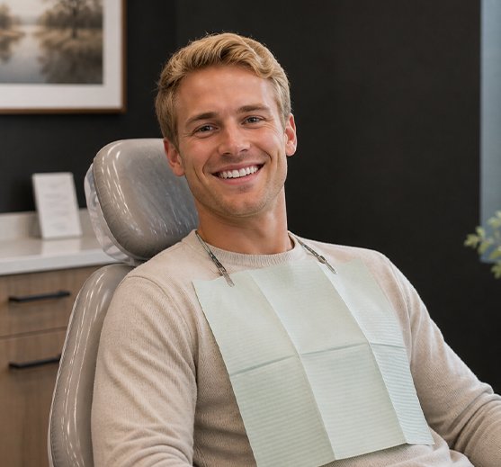 A smiling man wearing a dental bib sits in a dentist chair in a modern treatment room.