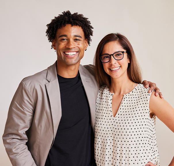 A smiling man and woman pose together, with the man wearing a gray blazer over a black shirt and the woman in a polka dot blouse.