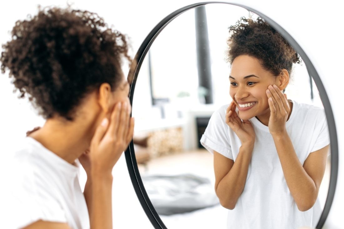 A young woman smiles and touches her face while looking into a mirror in a bright, modern room.