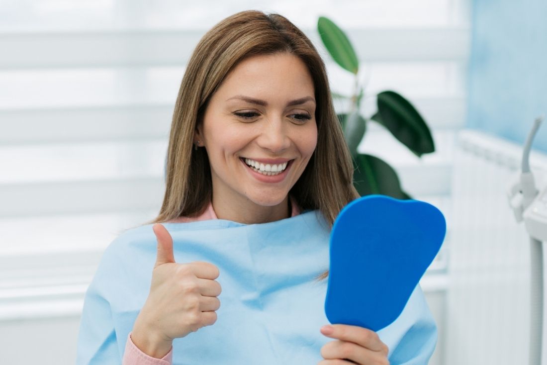 A smiling woman in a dental bib holds a blue mirror and gives a thumbs-up in a dental office setting.