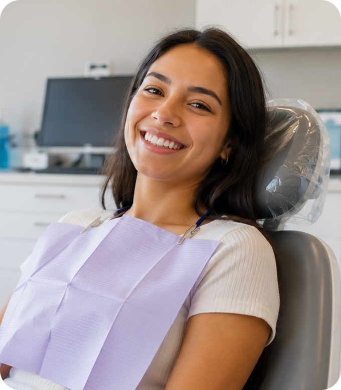 A patient smiling in a dental chair, wearing a purple bib, in a dentist's office with equipment in the background.