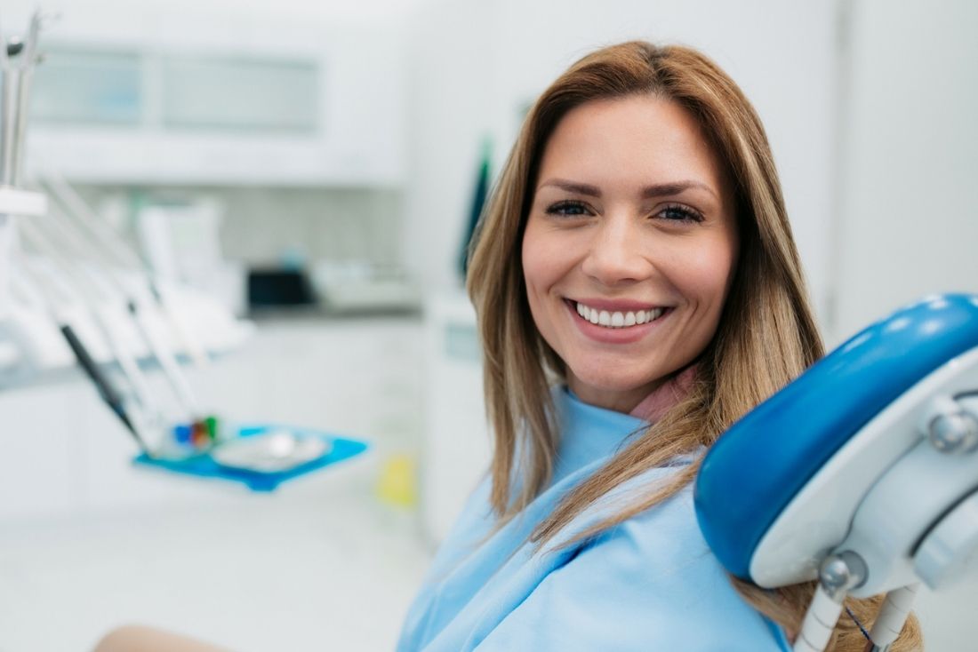 A patient smiling in a dental chair wearing a blue dental bib in a bright, modern dentist office.