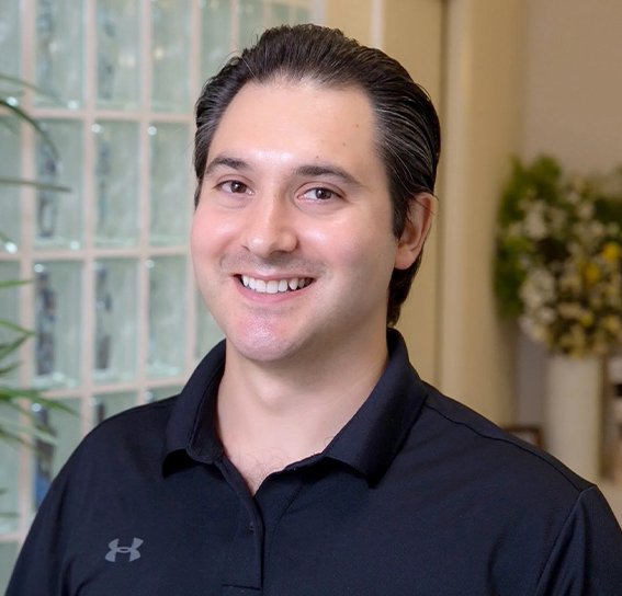 A smiling dentist in a black polo shirt stands in a dental office with glass block windows and greenery in the background.