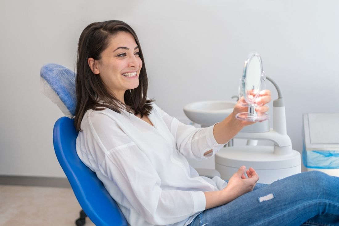 A smiling woman in a dental chair holding a small mirror, reviewing her teeth in a modern dentist office.