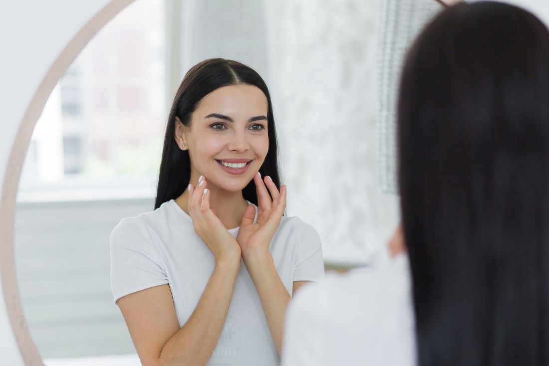 A young woman smiling at her reflection in a mirror, gently touching her face.
