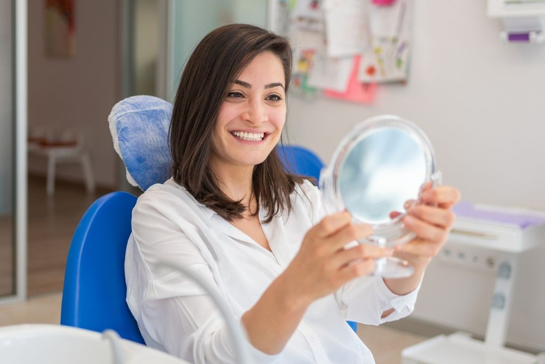 A woman smiles while holding a mirror, sitting in a dental treatment room.